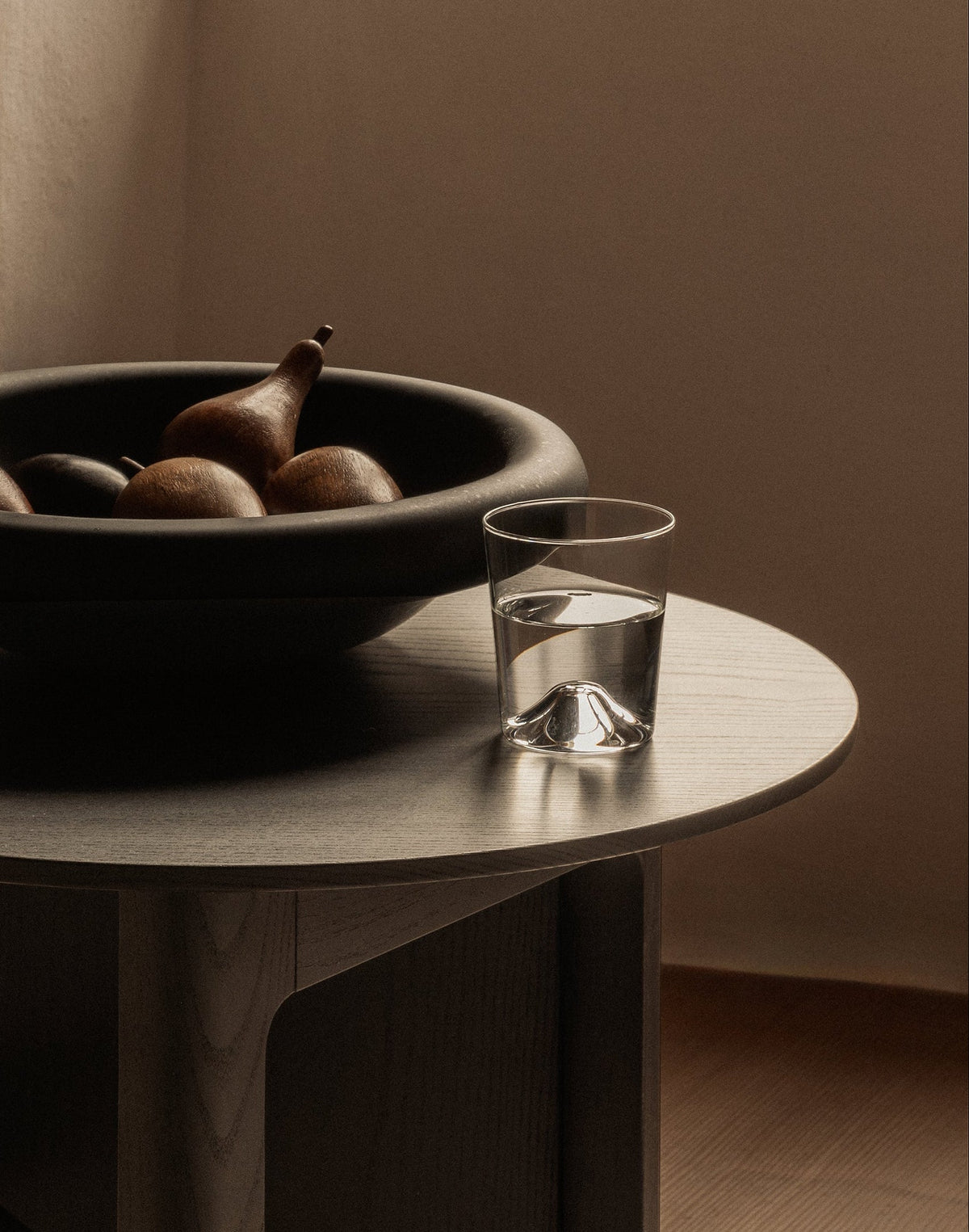 The Limmat Side Table in a warm, minimalist room, styled with a black bowl of fruit and a glass of water, next to a beige sofa.